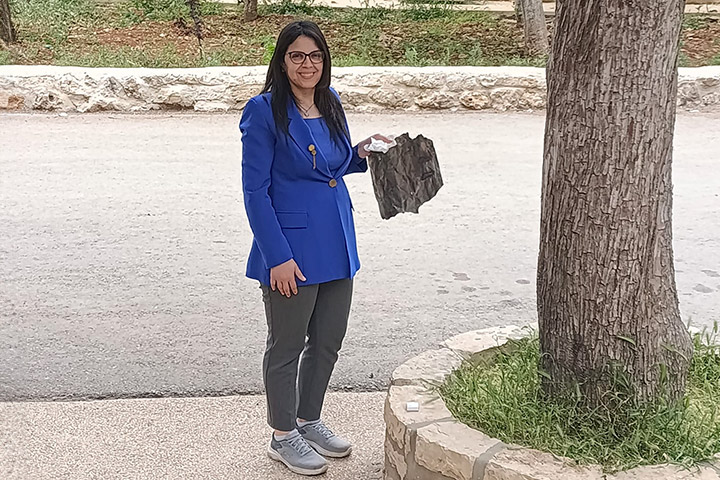 Laura Bishara, eine junge Frau mit langen dunklen Haaren, trägt eine Brille, ein blaues Sakko und Turnschuhe. Die palästinensische Schulleiterin von Talitha Kumi hält auf dem Schulgelände ein Raketentrümmerteil in der linken Hand, ein zerbeultes Blech.