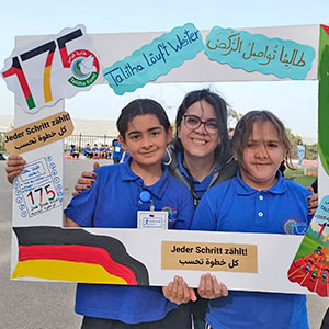 Bei einem Spenden-Marathon-Lauf in Talitha Kumi schauen Co-Schulleiterin Laura Bishara, eine junge Frau mit dunklen Haaren, und zwei Schülerinnen in blauen Shirts mit Schullogo durch einen selbstgebastelten Bilderrahmen aus Pappe. Auf ihm sind u.a. die Zahl 175 und eine Deutschlandfahne aufgeklebt.
