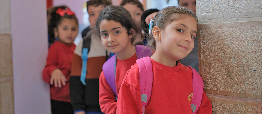 Einige Kita-Kinder kommen aus ihrem Klassenraum. Sie tragen rote Sweatshirts. Ein Mädchen im Vordergrund lächelt.