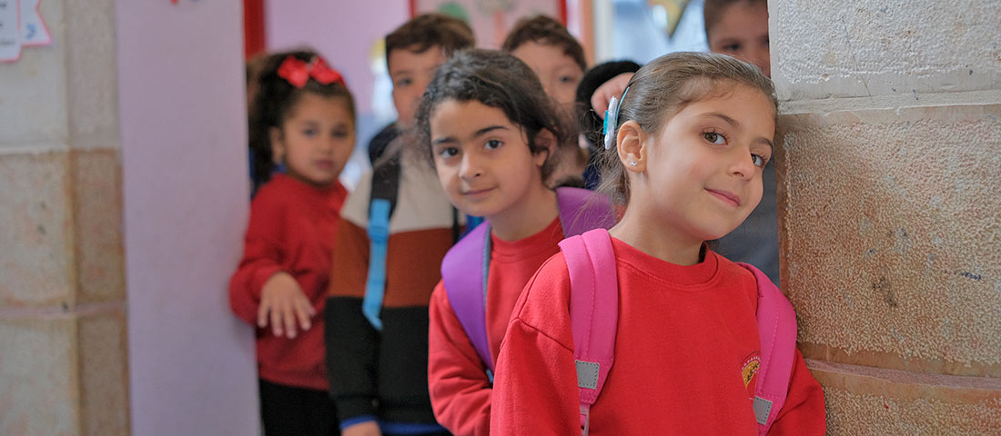 Einige Kita-Kinder kommen aus ihrem Klassenraum. Sie tragen rote Sweatshirts. Ein Mädchen im Vordergrund lächelt.