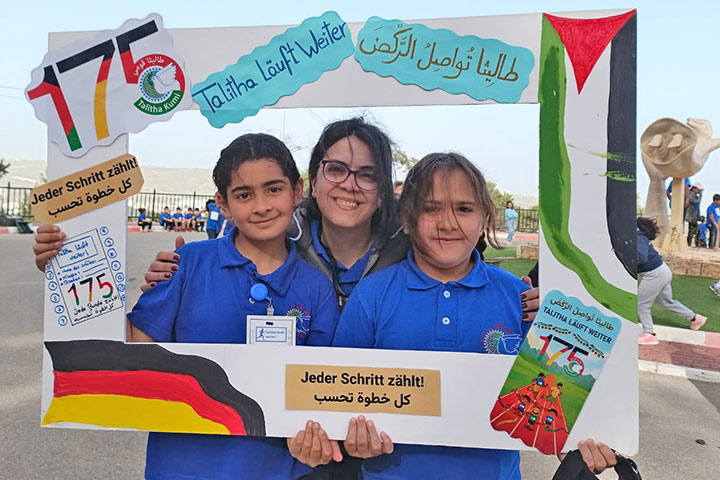 Bei einem Spenden-Marathon-Lauf in Talitha Kumi schauen Co-Schulleiterin Laura Bishara, eine junge Frau mit dunklen Haaren, und zwei Schülerinnen in blauen Shirts mit Schullogo durch einen selbstgebastelten Bilderrahmen aus Pappe. Auf ihm sind u.a. die Zahl 175 und eine Deutschlandfahne aufgeklebt.